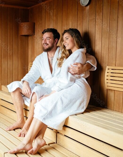 Man en vrouw samen in een Finse sauna, beiden in witte badjassen en glimlachend, terwijl de man zijn arm om de vrouw heen heeft in de binnen sauna