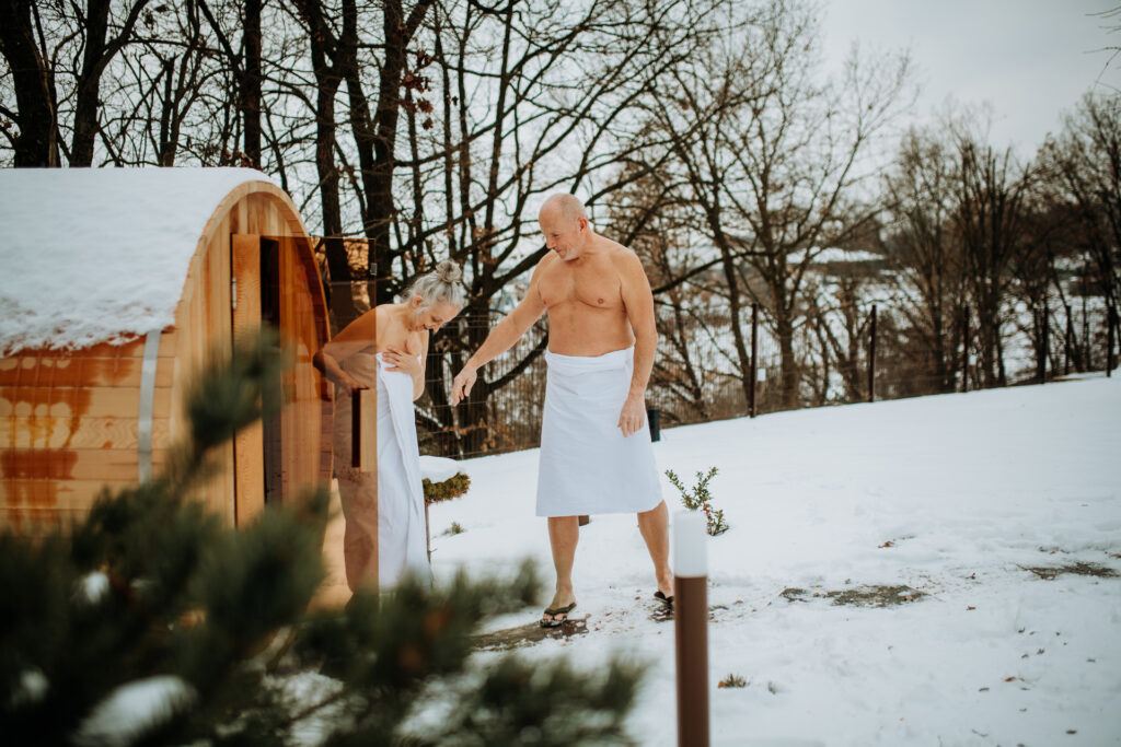 een man en vrouw in een witte badjas in een besneeuwd landschap stappen uit een barrelsauna