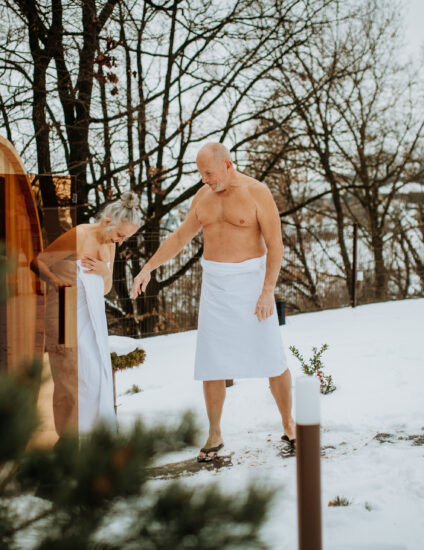 een man en vrouw in een witte badjas in een besneeuwd landschap stappen uit een barrelsauna