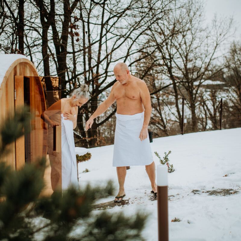 een man en vrouw in een witte badjas in een besneeuwd landschap stappen uit een barrelsauna