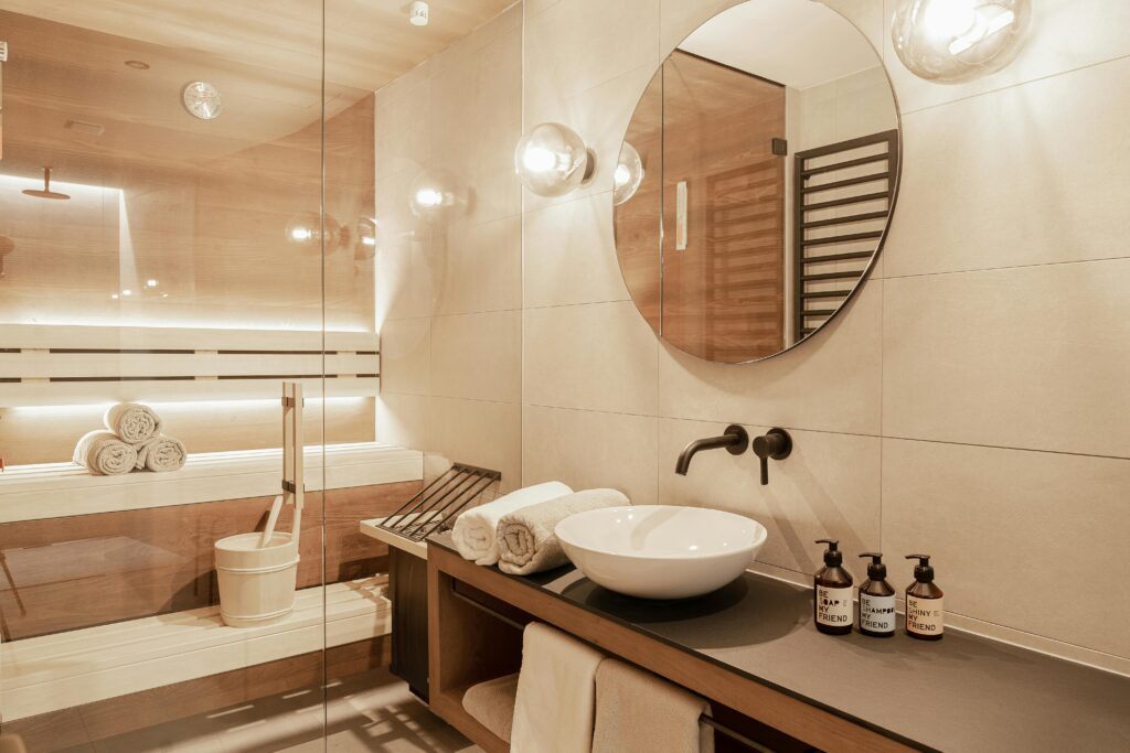 Een wit-beige badkamer met een grote ronde spiegel. In de badkamer staat een sauna waarin 3 handdoeken opgerold liggen