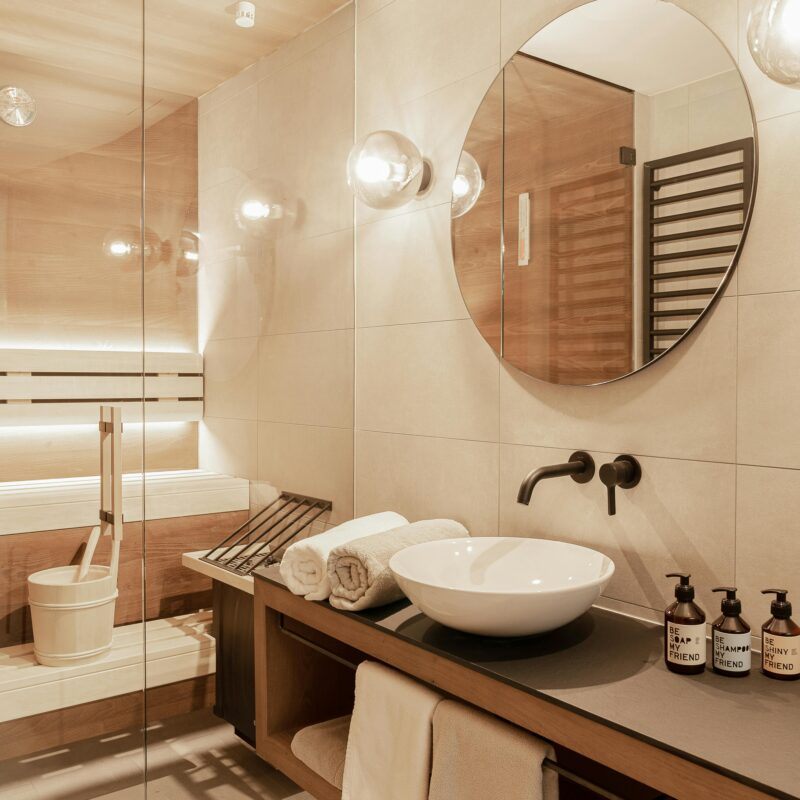 Een wit-beige badkamer met een grote ronde spiegel. In de badkamer staat een sauna waarin 3 handdoeken opgerold liggen