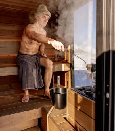 Een man in een sauna. Hij doet water op de hete kolen waardoor er stoom vrij komt. De sauna is een cedar hout combi sauna. De man draagt een grijze sauna hoed met de voordelen van dien.