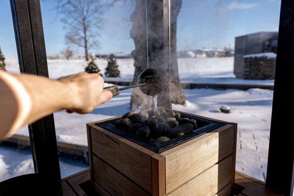 Op de afbeelding zie je een arm die water op heette sauna kolen gooit waardoor er stoom vrij komt. Op de achtergrond zie je een sneeuwlandschap