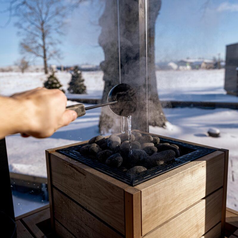 Op de afbeelding zie je een arm die water op heette sauna kolen gooit waardoor er stoom vrij komt. Op de achtergrond zie je een sneeuwlandschap
