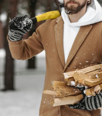 Man in een bruine mantal jas met daaronder een witte trui. Hij heeft houd in zijn recher hand en in zijn linker had een gele zaag om hout te hakken. Hij loopt in een koude besneeuwde bosrijke omgeving