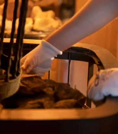 Ingezoomde foto van een hand met hittebestendige handschoenen die water op de saunastenen giet in een Finse sauna om de ruimte te verwarmen.