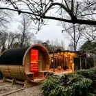 Barrelsauna in een herfstlandschap met warm rood sfeerlicht aan de binnenzijde en een gezellige overkapping met lampjes op de achtergrond