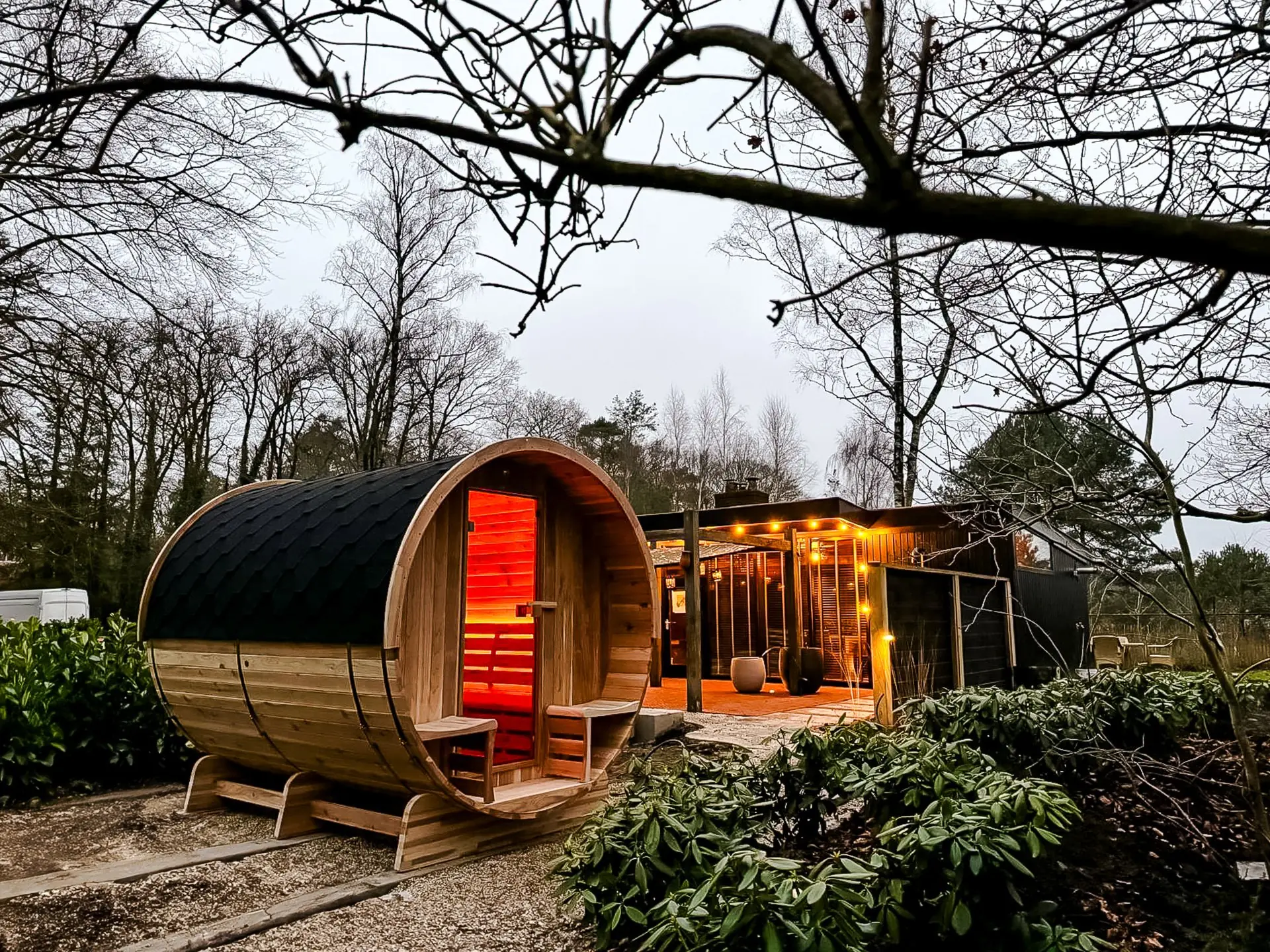 Barrelsauna in een herfstlandschap met warm rood sfeerlicht aan de binnenzijde en een gezellige overkapping met lampjes op de achtergrond