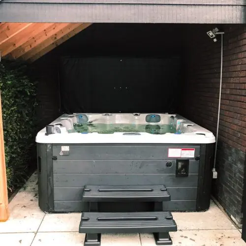 Jacuzzi spa met zwarte rand en zwart trappetje, geplaatst onder een afdak aan de zijkant van een huis. Een lampje brandt, hoewel de foto overdag is genomen