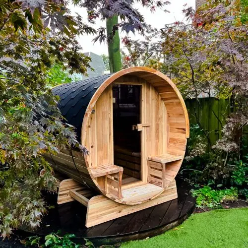 Barrel sauna in een tuin met veel groen en een boom in beeld