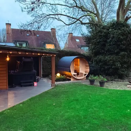 Groene tuin met een afdak achterin en een barrelsauna ernaast, waarbij de sfeerverlichting van de sauna brandt